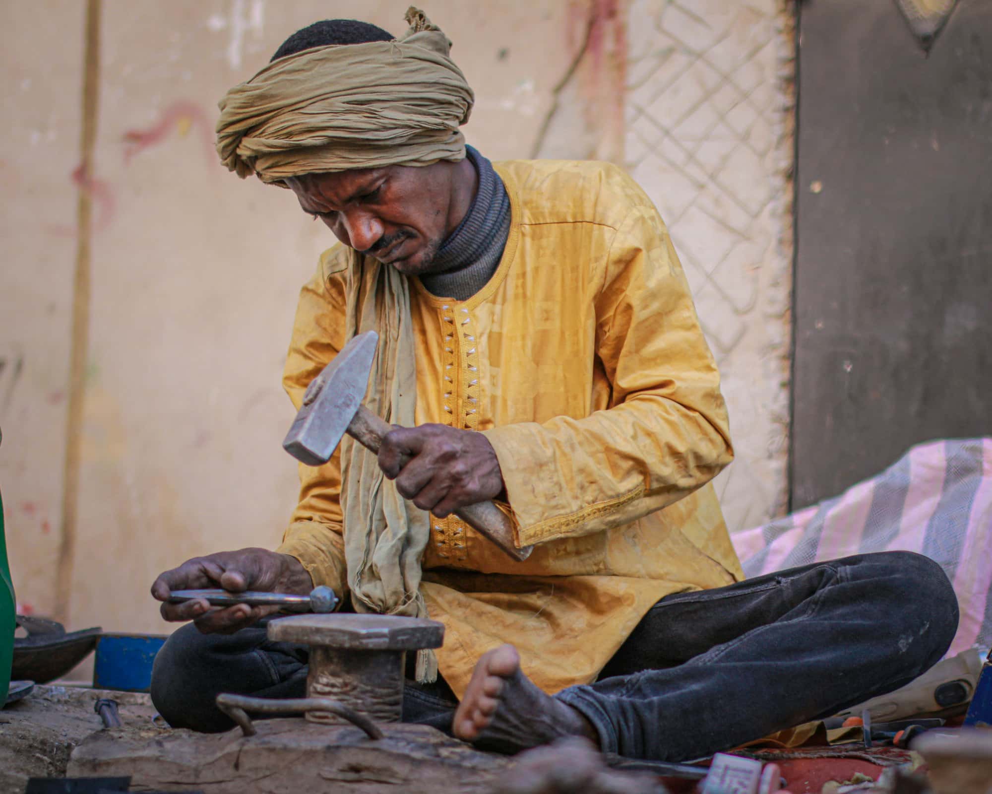 A craftsman sitting at a low anvil, hammering a punch into metal